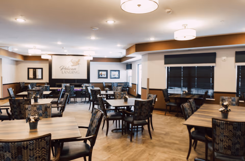 A spacious dining room with multiple wooden tables and patterned cushioned chairs arranged neatly. The room has warm lighting from ceiling fixtures and windows with dark blinds. A sign on the far wall reads 'Pelican Landing' with a pelican logo above it.