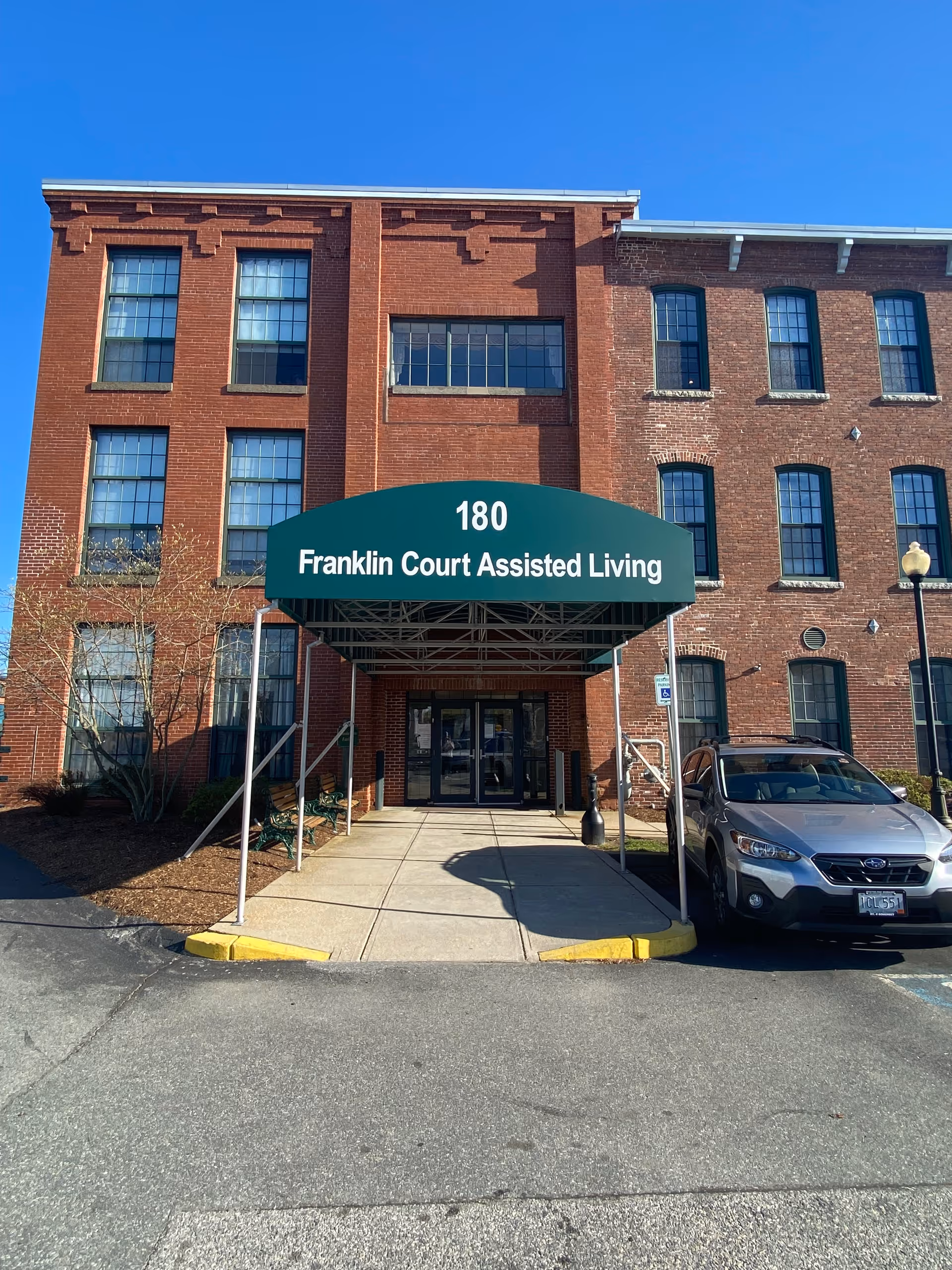 Front entrance of a brick building with a green awning that reads '180 Franklin Court Assisted Living'. There is a sidewalk leading to double glass doors, a parked car to the right, and a clear blue sky above.
