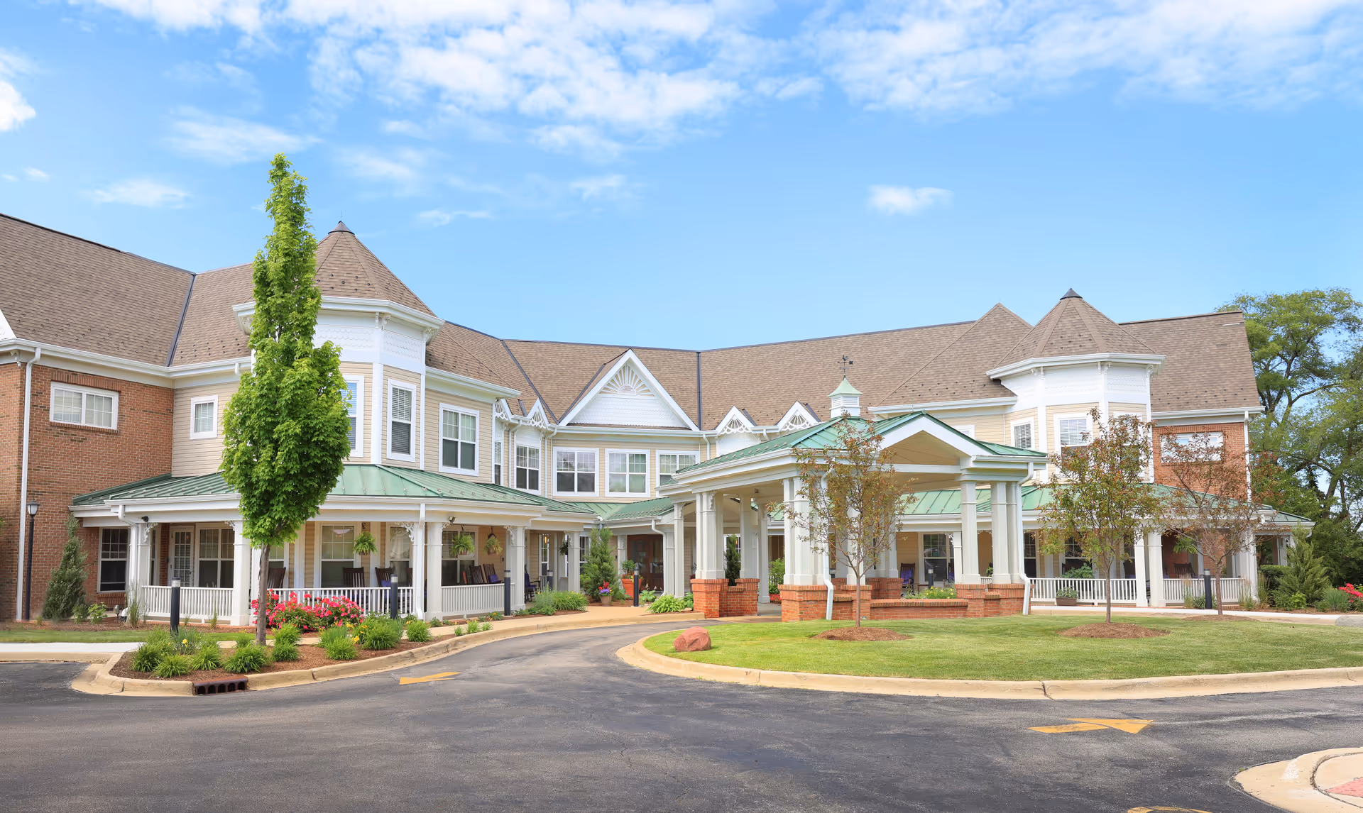 Front exterior view of a senior living facility building with a covered entrance, manicured lawn, trees, and a clear blue sky.