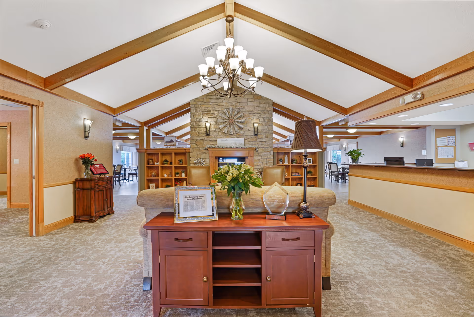 Spacious senior living lobby with vaulted wood‑beamed ceiling, central stone fireplace, reception desk and seating area.