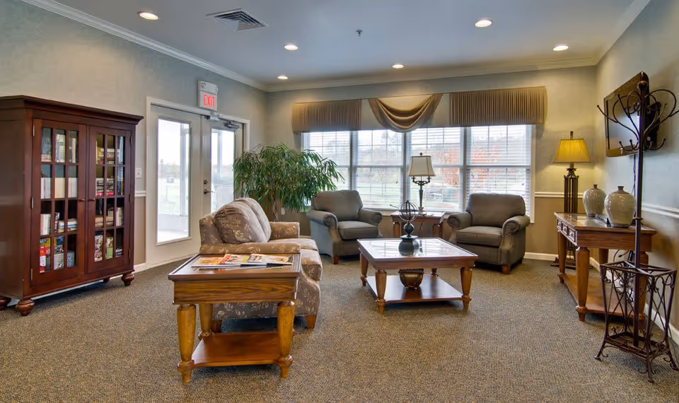 A cozy senior living facility common area with two armchairs and a patterned sofa arranged around a wooden coffee table. There is a wooden side table with magazines, a tall wooden cabinet with glass doors filled with books and games, a large window with beige curtains letting in natural light, a potted plant, a floor lamp, and a coat rack near the entrance door.