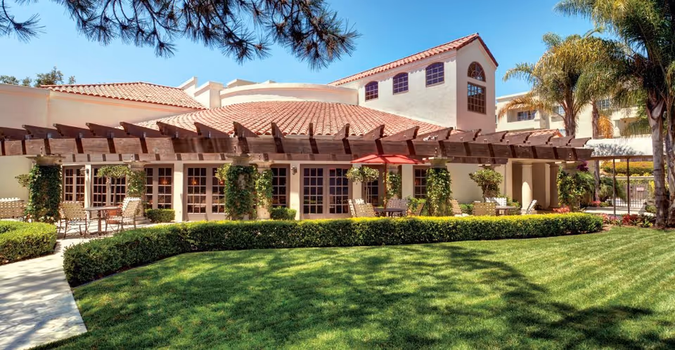 Outdoor view of a senior living facility with a well-maintained lawn, trimmed hedges, and a building featuring a tiled roof and large windows. There is a wooden pergola with climbing plants and outdoor seating including tables and chairs with an umbrella. Palm trees and other greenery surround the area under a clear blue sky.
