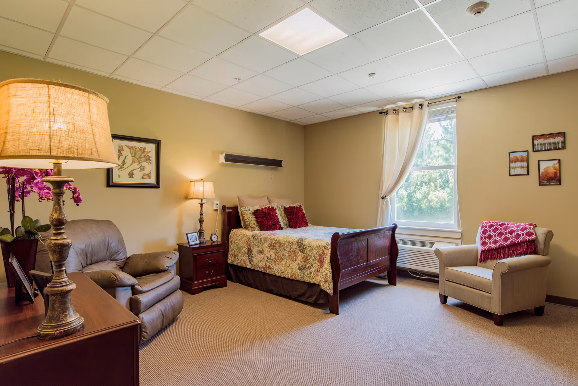 A cozy bedroom in a senior living facility with a wooden bed adorned with floral bedding and red pillows, a beige armchair with a red throw blanket, a brown recliner chair, a wooden nightstand with a lamp and small decor items, a window with white curtains letting in natural light, and framed artwork on the walls.