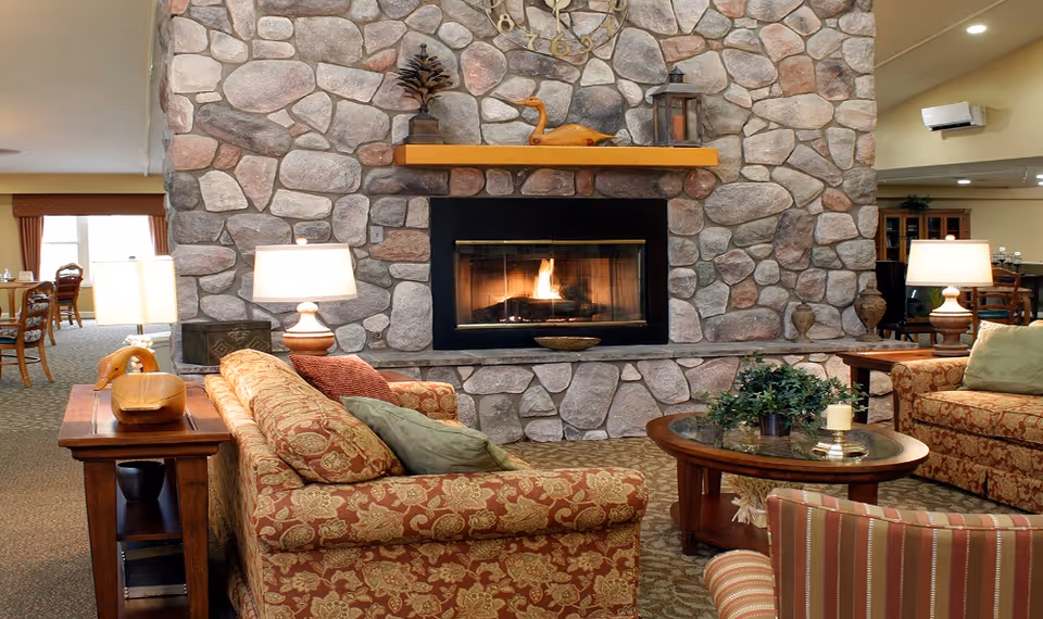 Cozy living room area with a stone fireplace in the center, a fire burning inside. The room features patterned sofas with cushions, a round glass coffee table with a plant and candle, wooden side tables with lamps, and decorative items on the fireplace mantel. In the background, there is a dining area with tables and chairs near windows with curtains.