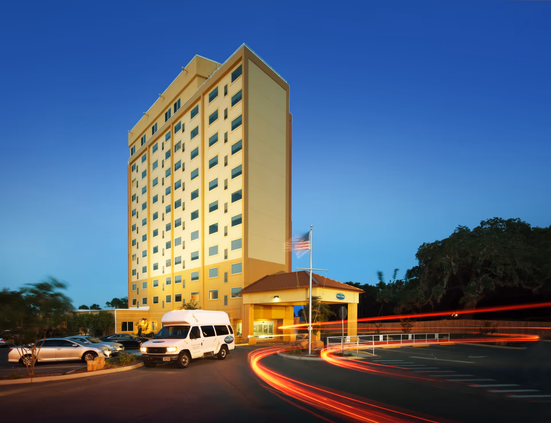 Exterior view of a tall, multi-story senior living facility building named Victoria Landing during dusk, with a shuttle van parked near the entrance and light trails from passing vehicles visible in the parking lot.