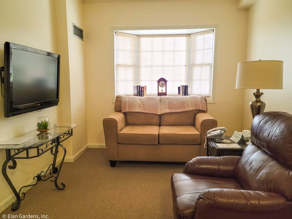 A cozy living room with a beige loveseat in front of a window with blinds, a brown leather armchair, a side table with a telephone and lamp, a wall-mounted flat screen TV, and a glass console table with a small plant.
