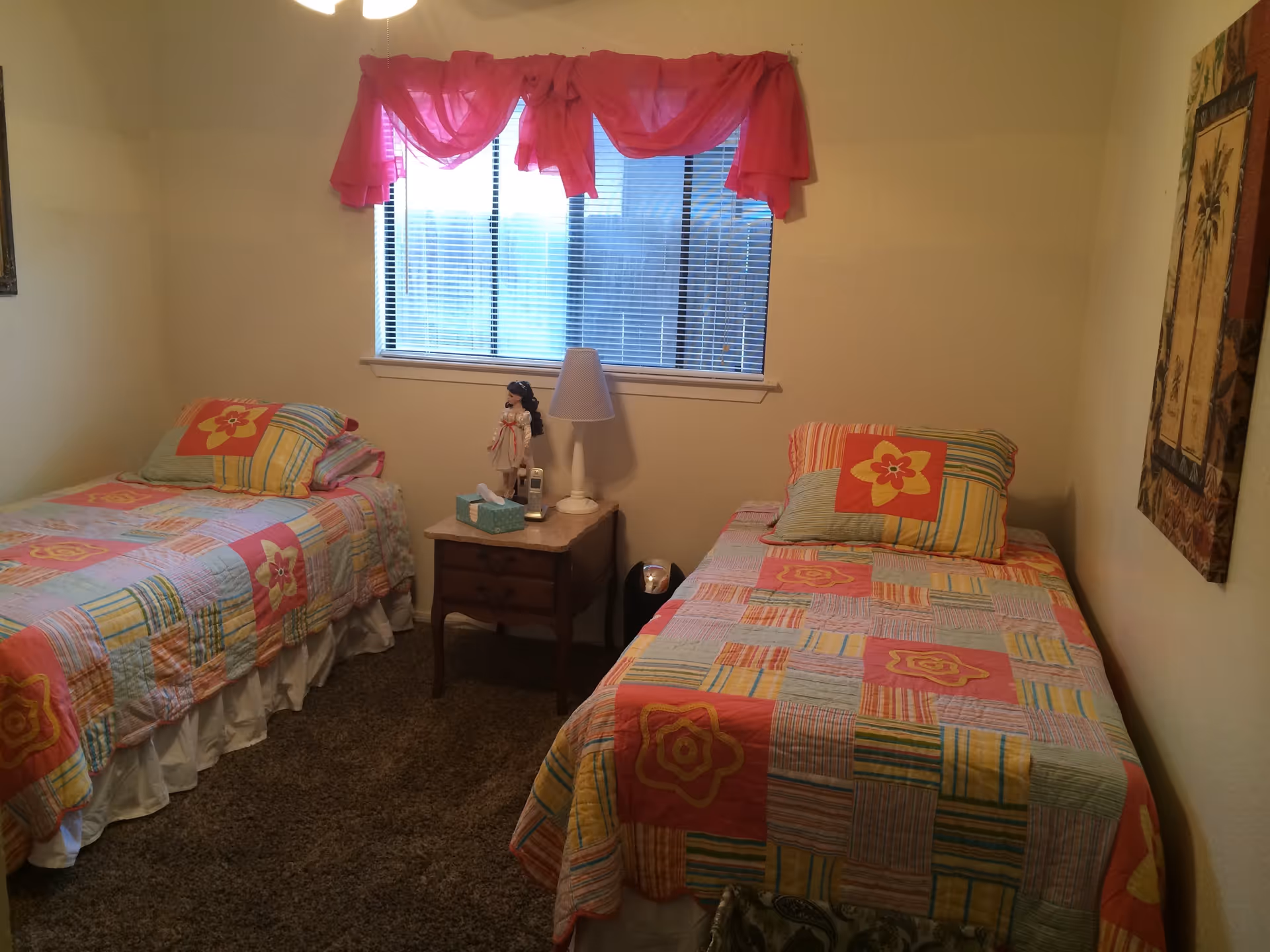 A small bedroom with two single beds placed against opposite walls. Each bed has colorful patchwork quilts with floral patterns and matching pillows. Between the beds is a wooden nightstand with a white lamp, a doll, a tissue box, and a cordless phone. A window with blinds and a pink valance is centered on the wall behind the nightstand. The room has beige walls and carpeted flooring.