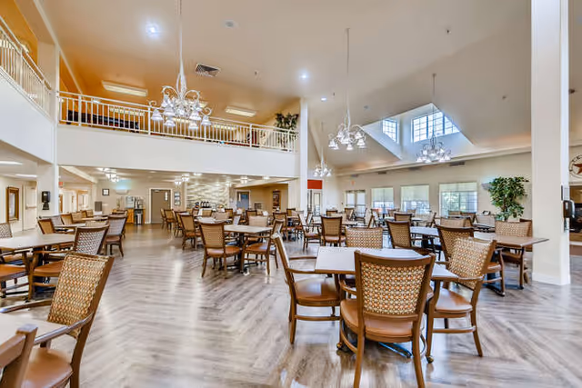 Spacious dining room in a senior living facility with multiple wooden tables and chairs arranged neatly. The room features high ceilings with large windows allowing natural light, chandeliers hanging from the ceiling, and a second-floor balcony overlooking the area. The flooring is light wood, and there are plants and decorative elements around the room.