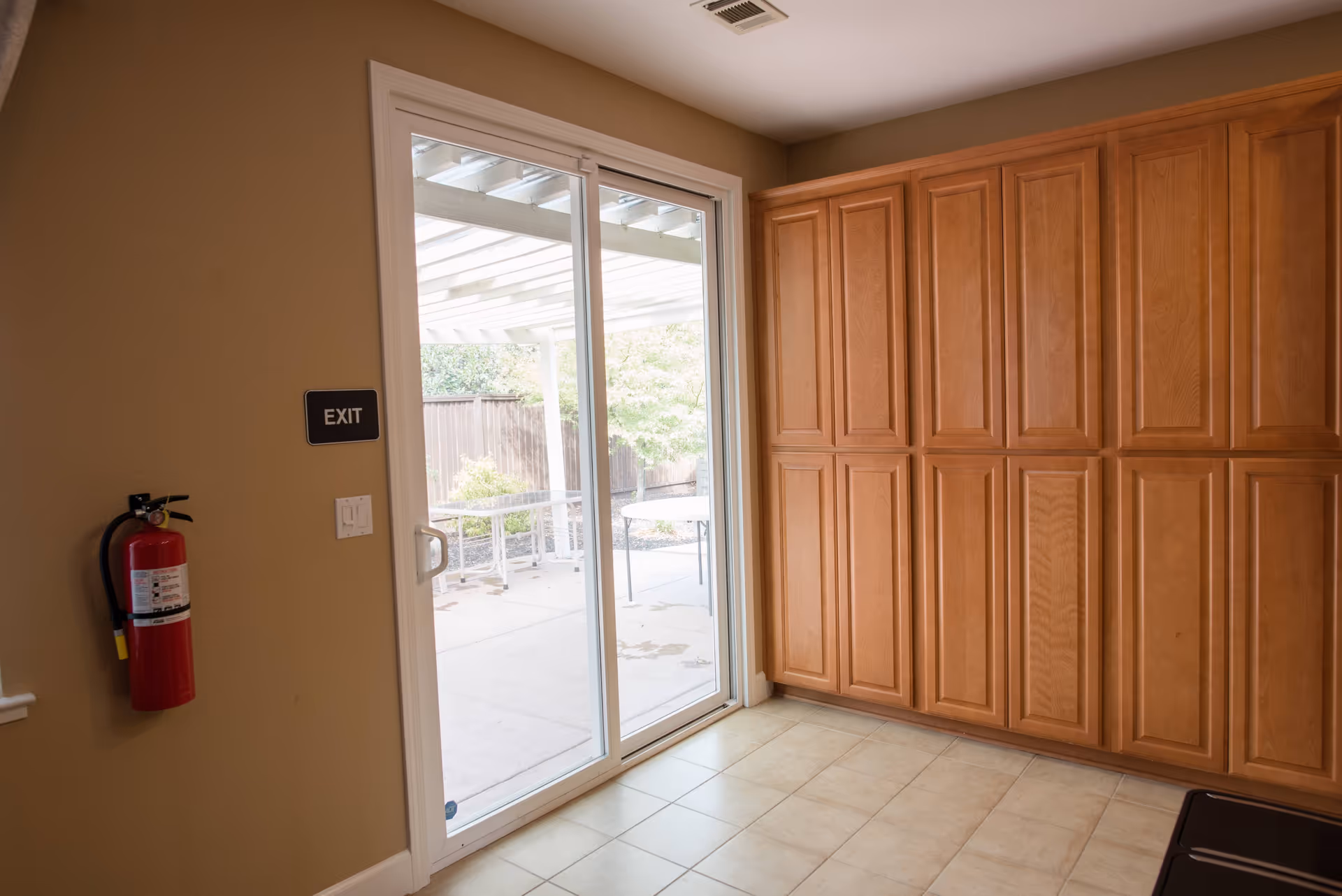 Interior view of a room with beige walls and tiled floor, featuring a large wooden cabinet on the right side and a sliding glass door leading to an outdoor patio area. A red fire extinguisher is mounted on the left wall below an exit sign.
