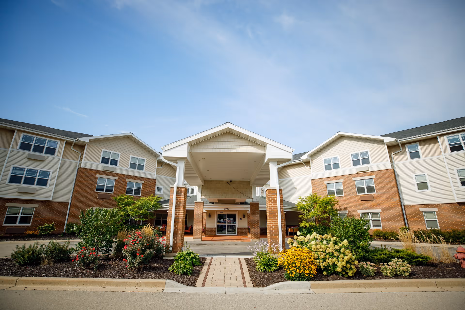 Front exterior view of a senior living facility building with a covered entrance, brick and beige siding, multiple windows, and landscaped flower beds with green shrubs and colorful flowers under a clear blue sky.