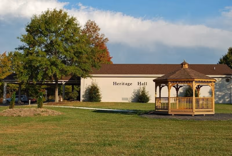 Exterior view of Heritage Hall building with a large green lawn, a wooden gazebo on the right, and a tree with green and autumn-colored leaves on the left under a partly cloudy sky.
