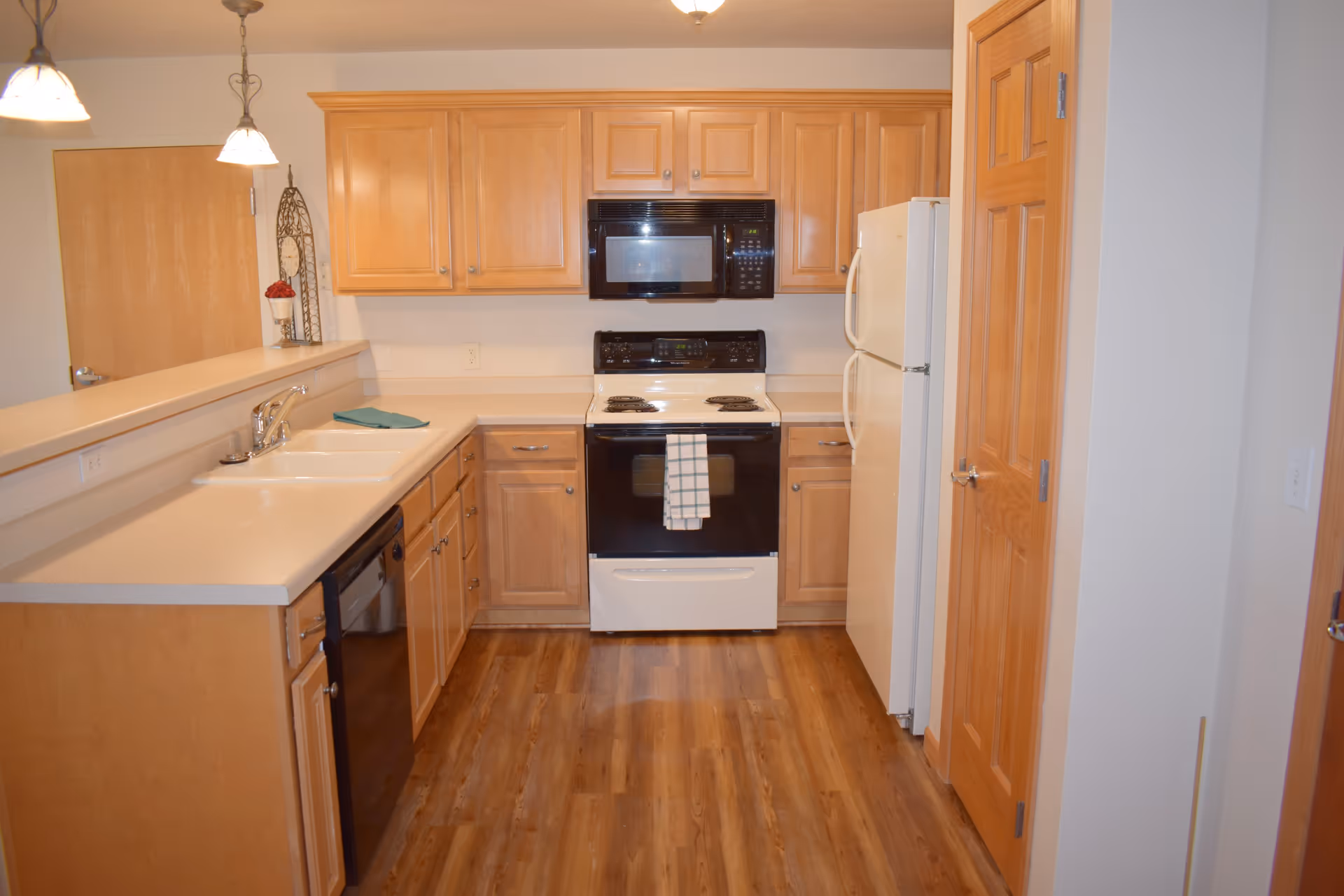 A kitchen with wooden cabinets, a white countertop, a white refrigerator, a black and white stove with a microwave above it, a dishwasher, and a double sink. The floor is wooden, and there are two pendant lights hanging from the ceiling. A towel is hanging on the oven handle.