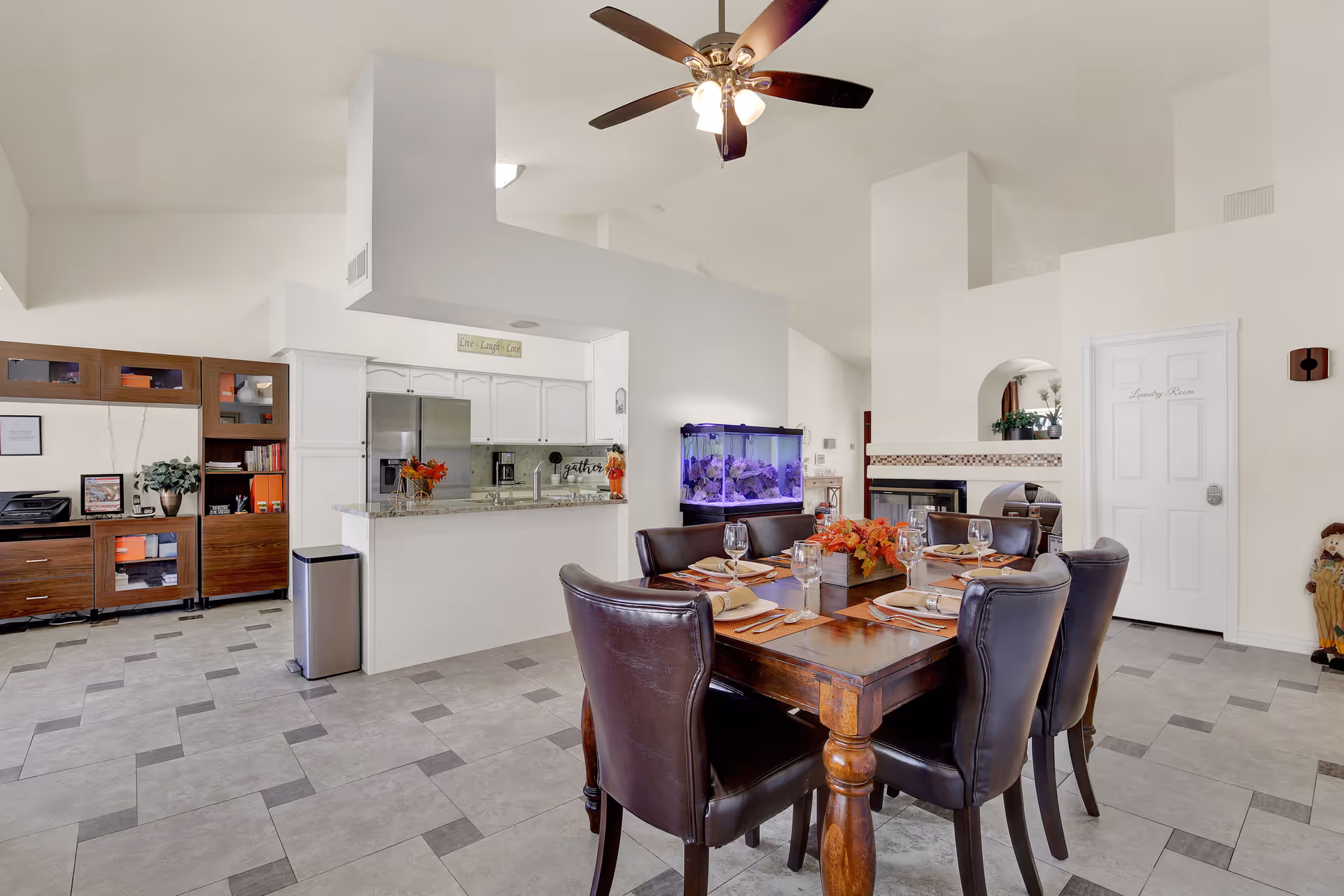 A bright and spacious dining area with a wooden dining table set for six with leather chairs. The room features a ceiling fan with lights, a large fish tank on a stand, and a kitchen area with white cabinets and granite countertops. There is a wooden cabinet with shelves and a printer on the left side, and a door labeled 'Laundry Room' on the right. The floor is tiled with a pattern of light and dark gray tiles.