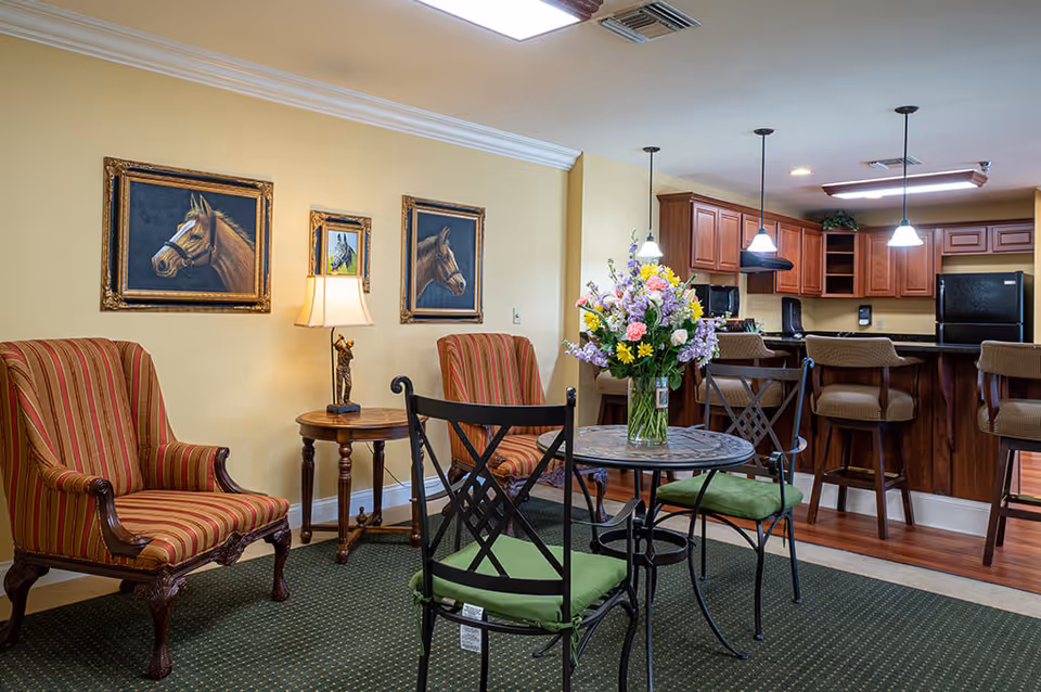 A cozy living area with two striped armchairs, a small round table with a lamp, and a round table with four chairs featuring green cushions. Behind the seating area is a kitchen with wooden cabinets, a black refrigerator, and three pendant lights hanging over a breakfast bar with four stools. The walls are decorated with three framed horse paintings.
