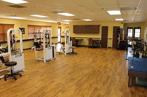 Spacious rehabilitation exercise room with fitness machines, therapy tables, and chairs on wood-look flooring.