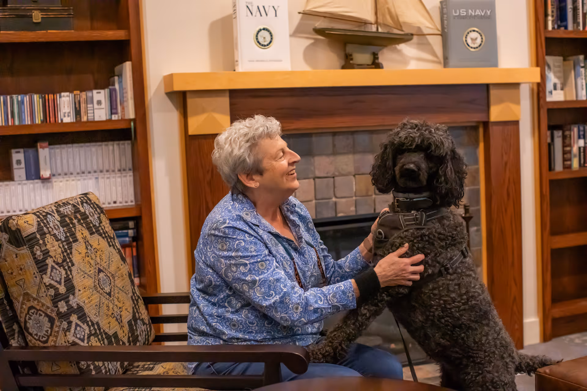 An elderly woman with short gray hair wearing a blue patterned shirt sits on a cushioned chair in a cozy room with bookshelves and a fireplace. She is smiling and petting a large black curly-haired dog that is standing on its hind legs with its front paws on her lap.
