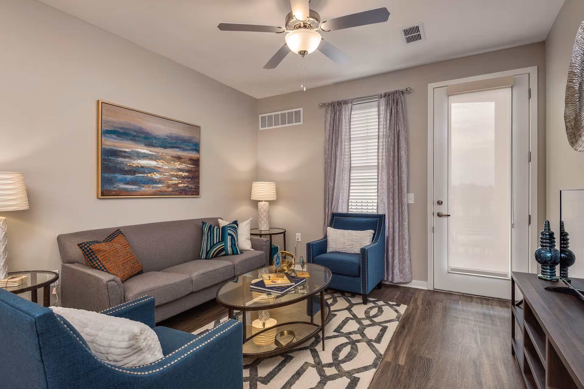 Stylish living room with a gray sofa, blue armchairs, a glass-top coffee table, wall art, and a door with window treatments.