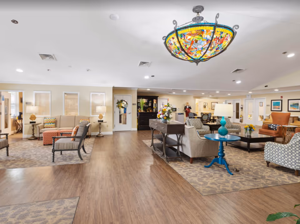 A spacious and well-lit senior living facility common area with comfortable seating arrangements including sofas, armchairs, and side tables. The room features a colorful stained glass chandelier hanging from the ceiling, wooden flooring, and decorative rugs. There are lamps on side tables, floral arrangements on a central table, and framed artwork on the walls. A person is visible in the background near a reception or service desk.