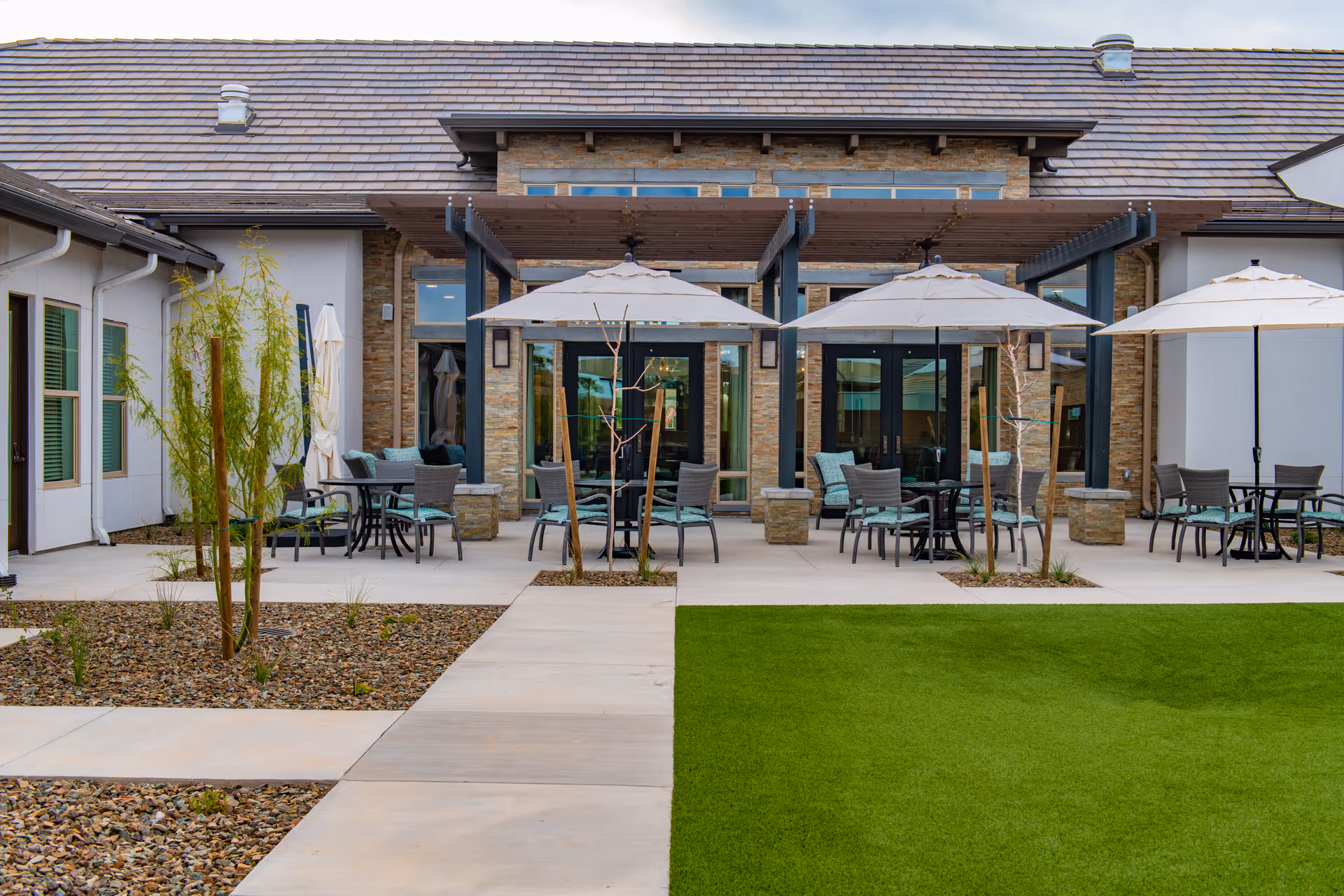 Outdoor patio area at Spring Gardens Senior Living Peoria featuring multiple tables with chairs and large umbrellas, surrounded by a stone and stucco building with large windows and a pergola overhead. The patio is adjacent to a green lawn and landscaped areas with small trees and rocks.