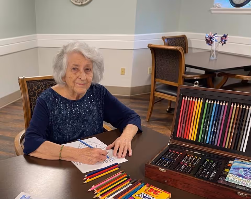 An elderly woman with white hair is sitting at a table in a senior living facility, coloring a drawing with colored pencils. On the table, there is a large open art set with various colored pencils, crayons, and drawing supplies. The room has wooden floors, light-colored walls, and several chairs and tables in the background.