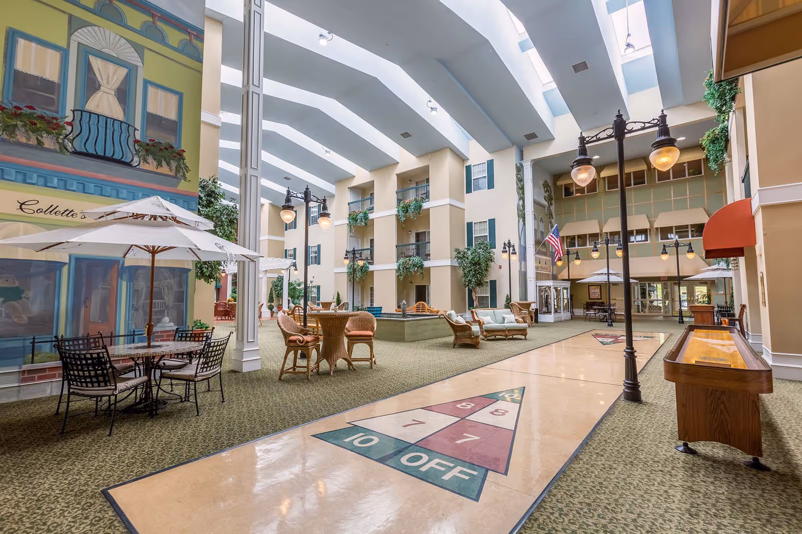 Indoor common area with high skylight ceiling, featuring shuffleboard courts on the floor, wicker chairs and tables with umbrellas, streetlamp-style lighting, and decorative plants. The walls have painted window and balcony designs, and there is an American flag near the entrance.