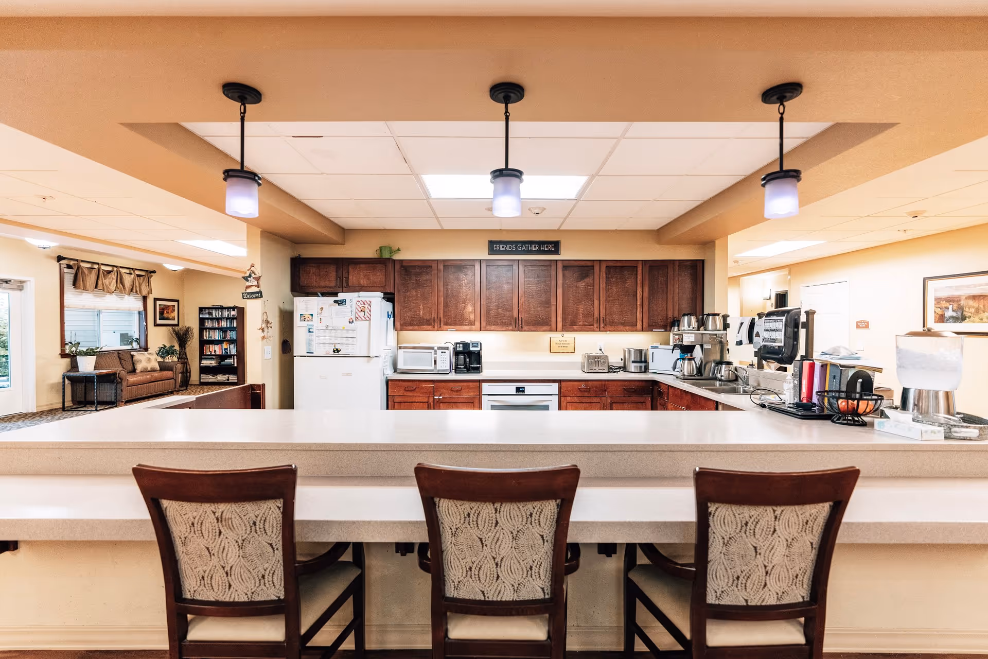 A bright and clean kitchen area in an assisted living facility with a long counter and three wooden chairs with patterned upholstery. The kitchen features wooden cabinets, a white refrigerator, microwave, coffee makers, and various kitchen appliances. Pendant lights hang from the ceiling above the counter. In the background, there is a seating area with a couch, a bookshelf, and a window with curtains.