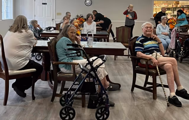 A group of elderly people sitting in a common room with wooden chairs and tables. Some individuals are using walkers or canes. The room has light-colored walls and flooring, with a clock on the wall and shelves stocked with various items in the background. A few people are engaged in conversation or looking towards the camera.