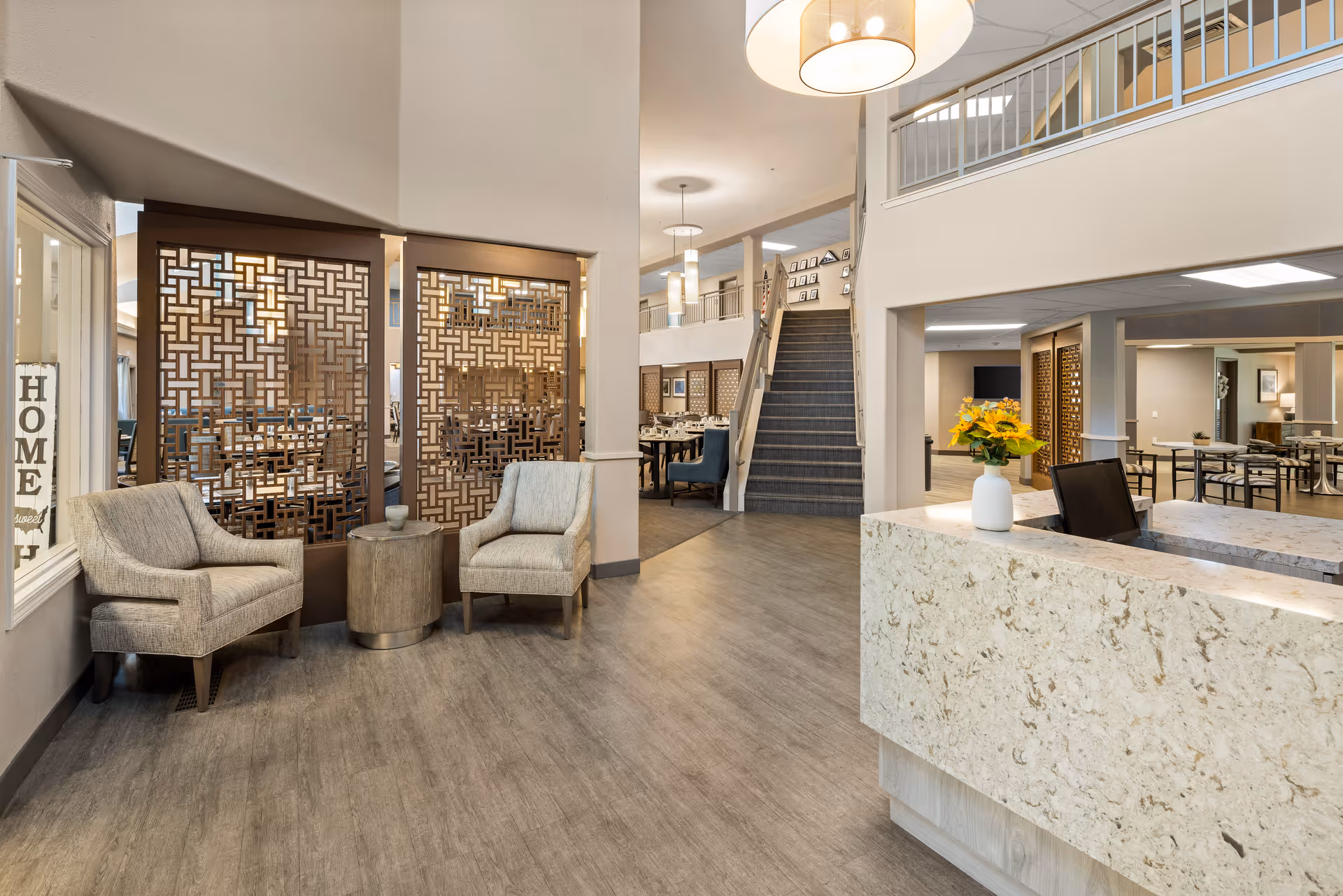 Interior view of a senior living community featuring a reception desk with a vase of yellow flowers, two beige armchairs with a small round table between them, decorative wooden room dividers, and a staircase leading to an upper floor. The space is well-lit with hanging ceiling lights and has a modern, welcoming design.
