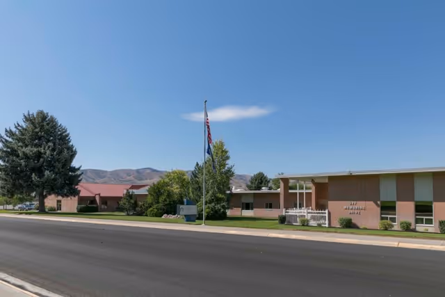 Low-rise brick care facility building with an American flag and landscaping along a street with mountains in the background.