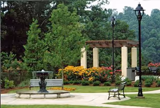Outdoor garden area with a small fountain in the center, surrounded by benches, lampposts, colorful flowers, and tall green trees with a pergola structure in the background.