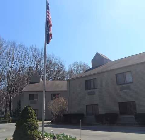Exterior view of a two-story building with beige siding and multiple windows, surrounded by leafless trees and shrubs. An American flag is flying on a flagpole in front of the building under a clear blue sky.