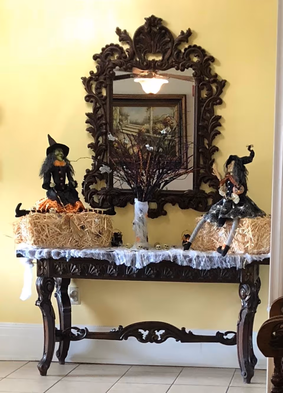 A decorative wooden table with intricate carvings against a yellow wall, topped with two Halloween witch dolls sitting on hay bales, a vase with branches, and small skull decorations. Above the table is an ornate framed mirror reflecting a ceiling fan and a framed painting.