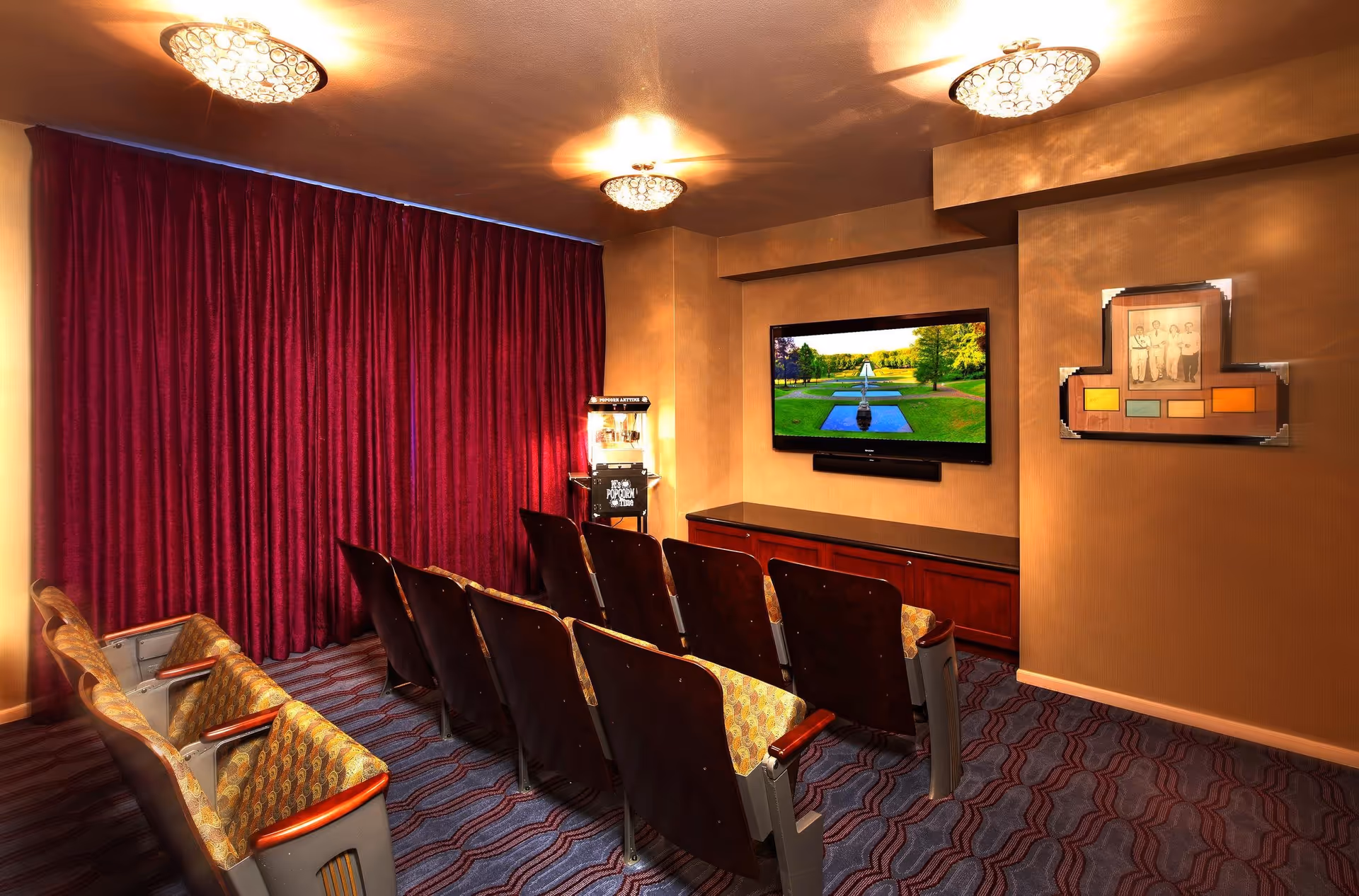 Small screening room with rows of theater seats facing a wall-mounted TV, a popcorn machine, and a closed red curtain.
