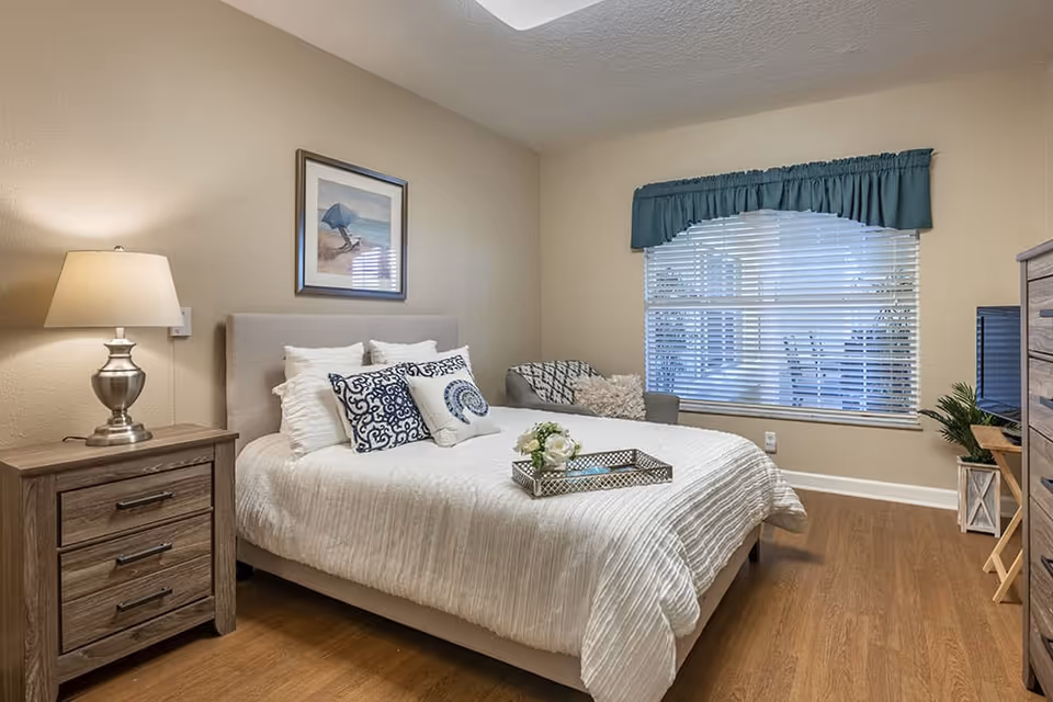 A cozy bedroom with a neatly made bed featuring white bedding and decorative pillows. There is a wooden nightstand with a lamp on the left side of the bed. A framed picture hangs on the wall above the bed. A large window with white blinds and a blue valance lets in natural light. A small armchair with cushions is placed near the window. On the right side, there is a wooden dresser with a TV on top and a small plant beside it. The floor is wooden.