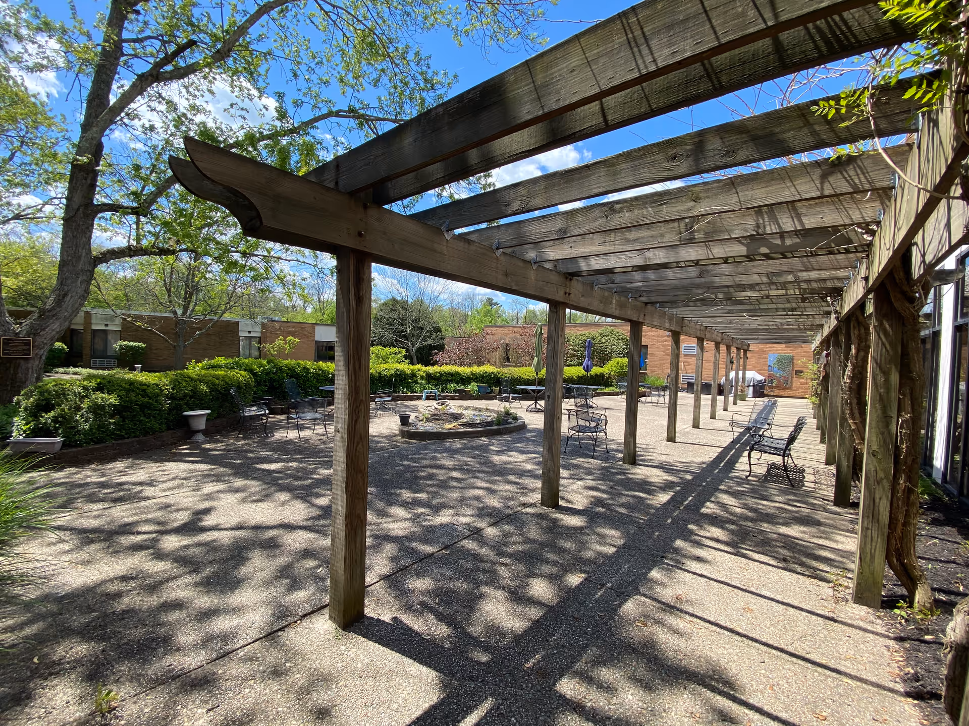 Outdoor courtyard area with a wooden pergola casting shadows on the paved ground. There are benches and metal chairs arranged around a circular stone planter with greenery. Surrounding the courtyard are bushes, trees, and brick buildings under a blue sky with some clouds.