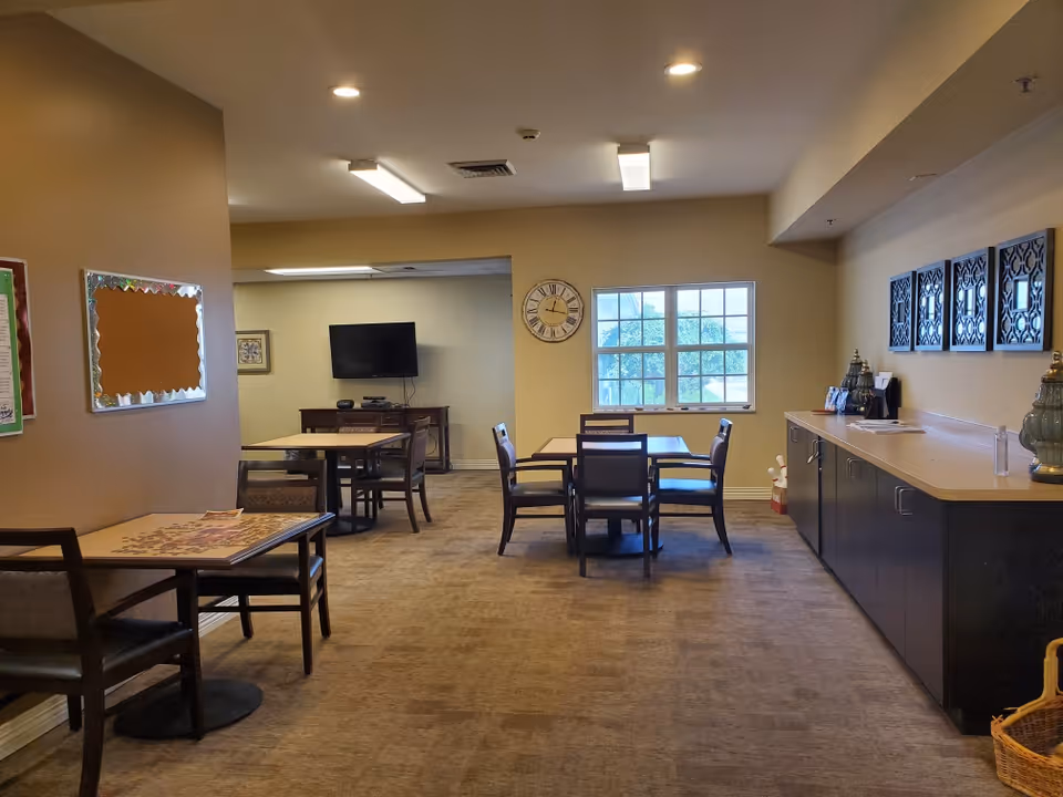 A senior living common room with multiple tables and chairs, a wall-mounted TV, a clock by a window, and a long counter with cabinets.