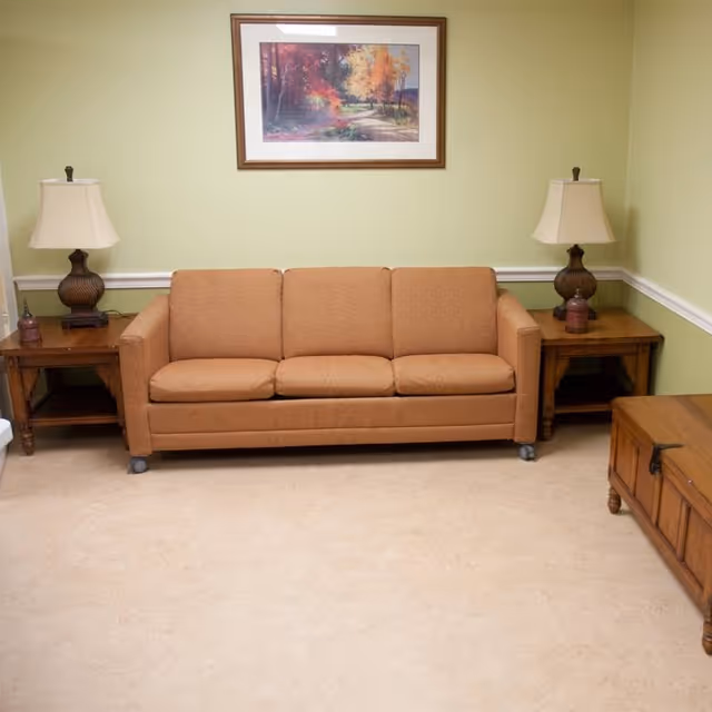 A small sitting area with an orange three-seat sofa flanked by wooden side tables and lamps beneath a framed landscape painting.
