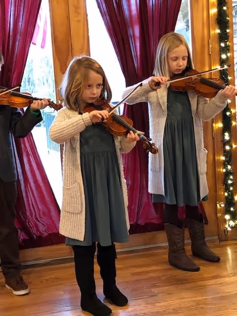 Two young girls standing indoors playing violins in front of a window with red curtains and holiday string lights on the wooden frame.