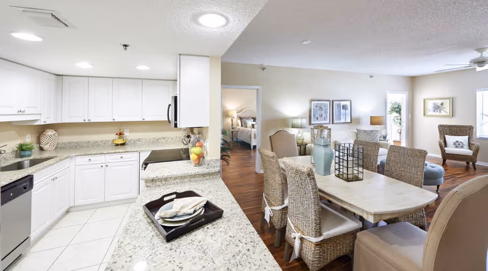 Bright and spacious open-concept kitchen and dining area with white cabinets, granite countertops, and wicker dining chairs around a rectangular table. The living room is visible in the background with a sofa, armchairs, framed artwork on the walls, and a ceiling fan. A bedroom can be seen through an open doorway.