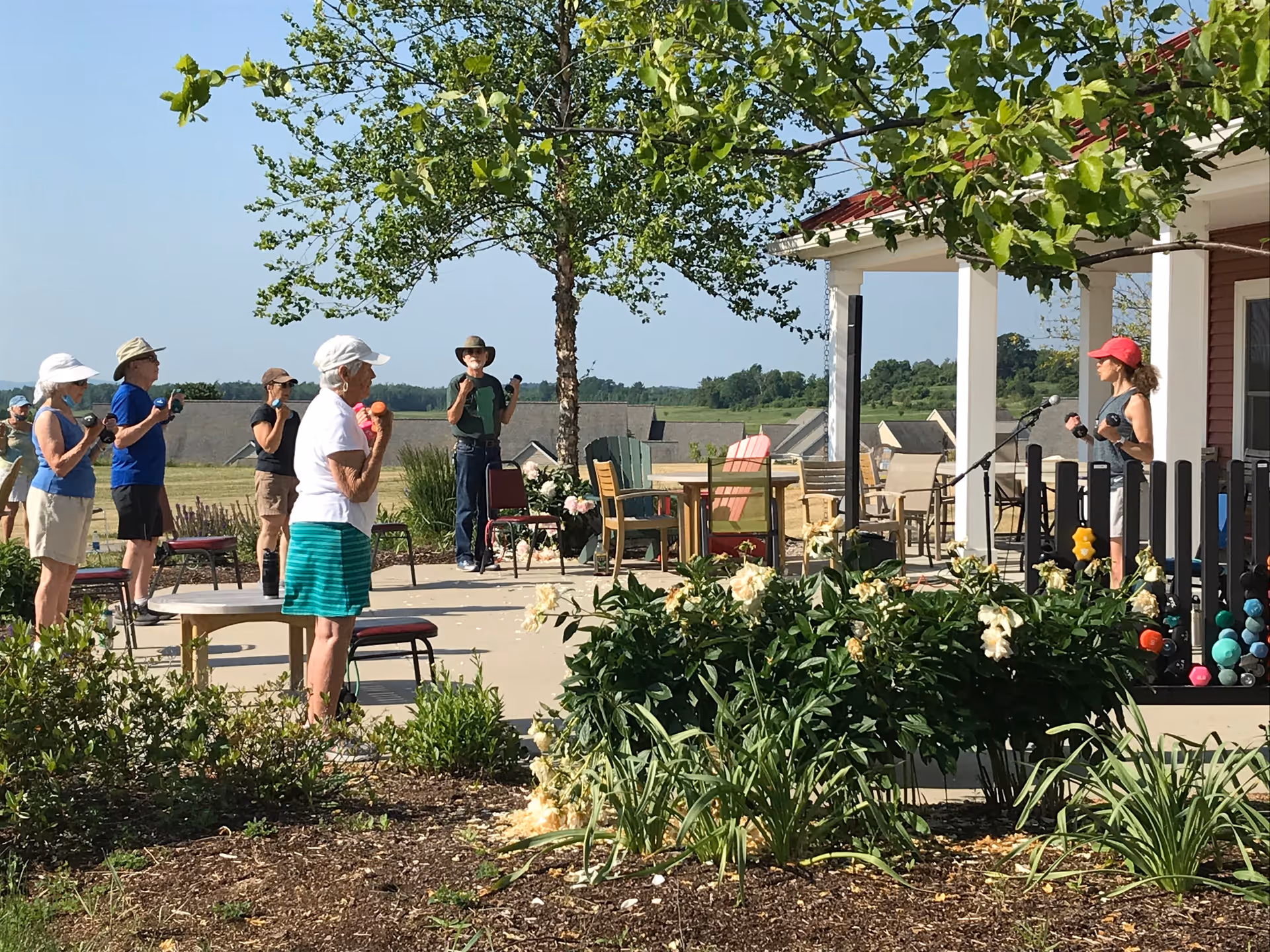 Seniors participating in an outdoor exercise class on a patio near a porch and garden beds.