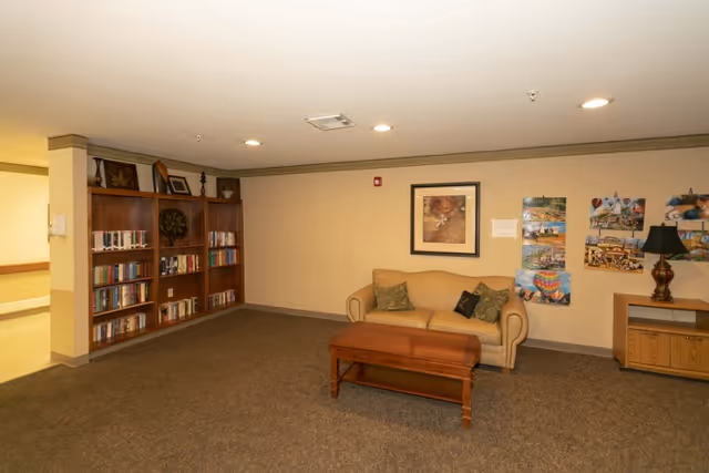 Small common room with a beige couch, wooden coffee table, bookshelf filled with books, framed artwork and a lamp.