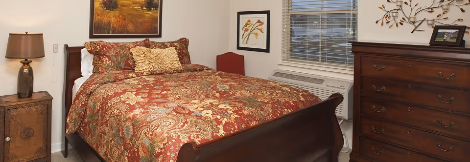 A cozy bedroom with a wooden sleigh bed covered in a red and gold floral patterned comforter and decorative pillows. There is a wooden nightstand with a lamp on the left side of the bed, a wooden dresser on the right side of the room, a window with blinds, and framed artwork on the walls.