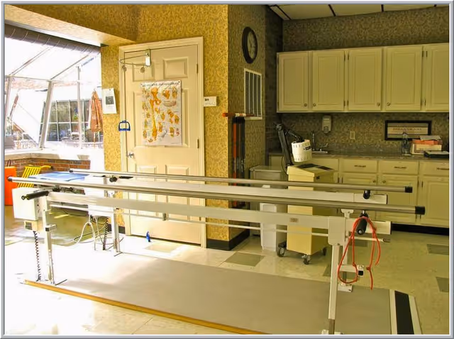 A sunlit interior therapy room with parallel walking bars, cabinets, and medical equipment.