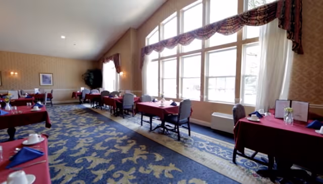 A spacious dining room with large windows letting in natural light. The room features tables covered with red tablecloths, each set with blue napkins, white cups, and small flower arrangements. The carpet has a blue and gold ornate pattern, and the walls are decorated with patterned wallpaper and framed artwork. There are elegant curtains above the windows and wall sconces providing additional lighting.