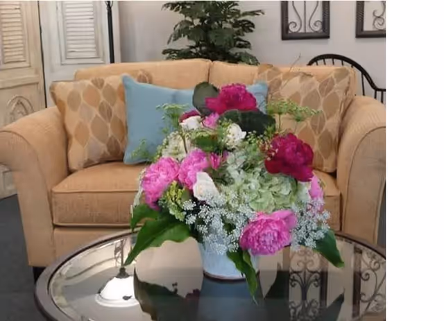 A beige couch with patterned cushions and a blue pillow behind a glass coffee table holding a large bouquet of pink, white, and green flowers. A green plant and framed wall decorations are visible in the background.