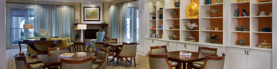 A well-lit living room area with multiple wooden tables and chairs arranged neatly. The room features large windows with curtains, a fireplace with a framed painting above it, and built-in shelves filled with decorative items and books. The carpet has a patterned design, and there are table lamps providing additional lighting.