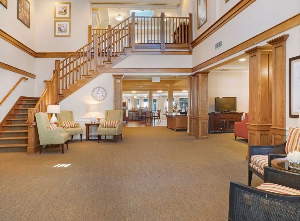 A spacious and well-lit common area in a senior living facility featuring a wooden staircase with a railing leading to an upper floor. The room has beige carpeting, wooden trim, and columns. There are several seating areas with armchairs and small tables, a wall clock, framed artwork, and a television on a wooden stand. The space opens into a dining area in the background.