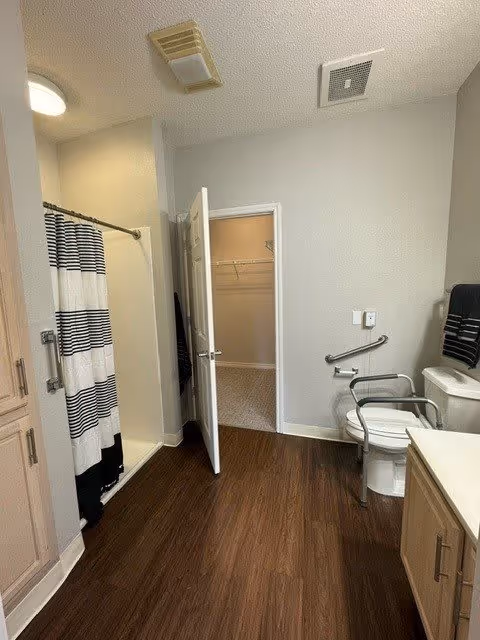 A bathroom with a shower area on the left featuring a black and white striped shower curtain. The floor is wooden, and there is a white toilet with safety rails on the right side. A towel rack with a black towel is mounted on the wall above the toilet. The bathroom door is open, revealing a walk-in closet with carpeted flooring. The walls are painted light gray, and there are ceiling vents and a light fixture visible.