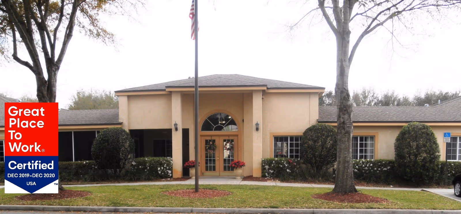 Exterior front view of a single-story beige building with a central entrance featuring double glass doors and an arched window above. There are two large trees and neatly trimmed bushes in front, with a flagpole displaying the American flag in the center. A parking area is visible to the right.