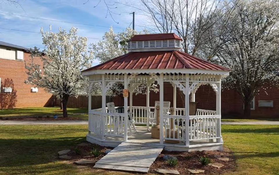 A white wooden gazebo with a red roof situated on a grassy area surrounded by blooming trees and a brick building in the background under a clear blue sky.